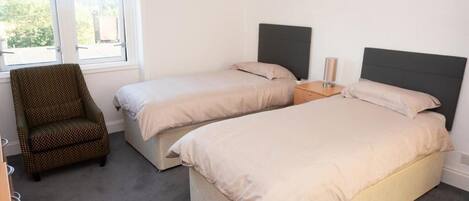 Un dormitorio con dos camas individuales, una silla, una mesita de noche y un ventanal.