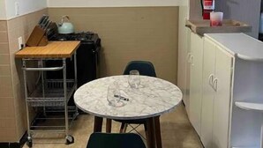 Una cocina pequeña con una mesa redonda, dos sillas, un carrito metálico y un armario blanco.