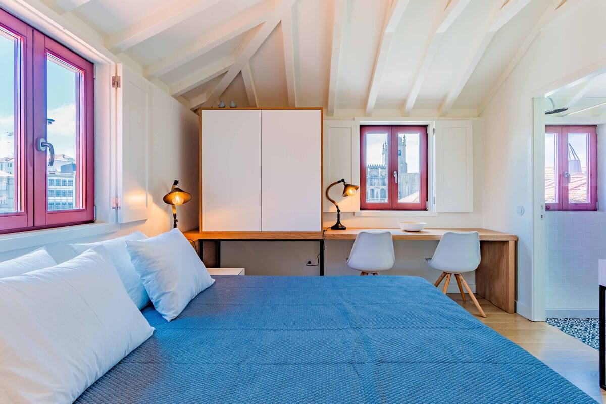 A bedroom with a large bed, a desk, and a chair. There are windows with pink frames and a view of buildings outside.