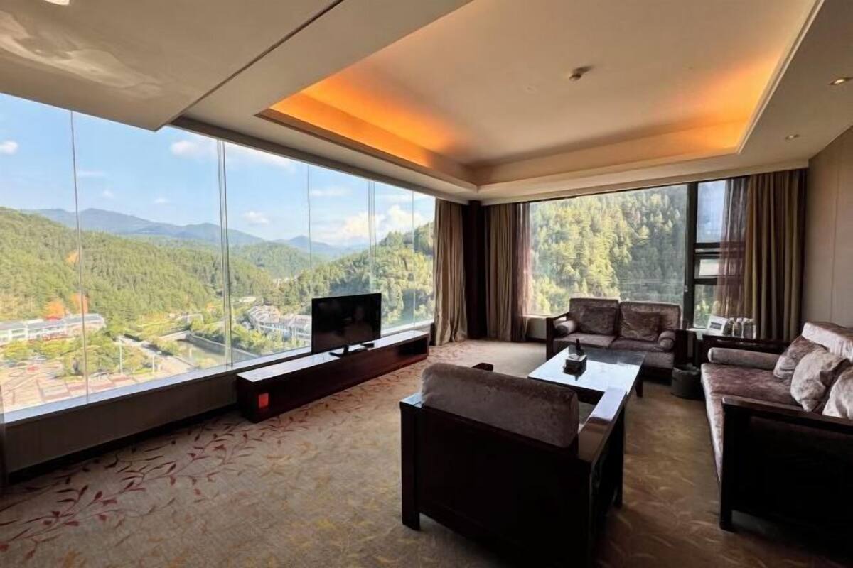 A modern hotel room with a large window offering a mountain view, a flat-screen TV, and a cozy seating area with a sofa and armchairs.