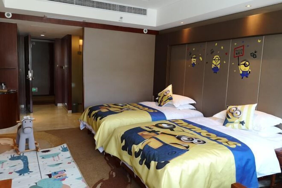 A hotel room with two beds, each covered with a Minions-themed duvet. There is a wooden floor, a small rug with cartoon characters, and a door leading to another room.