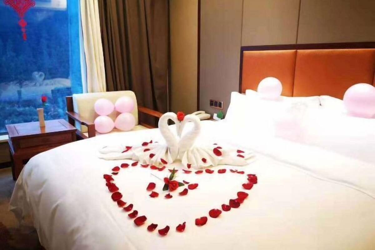 A hotel room with a neatly made bed, a heart-shaped arrangement of rose petals, and a swan-shaped centerpiece.