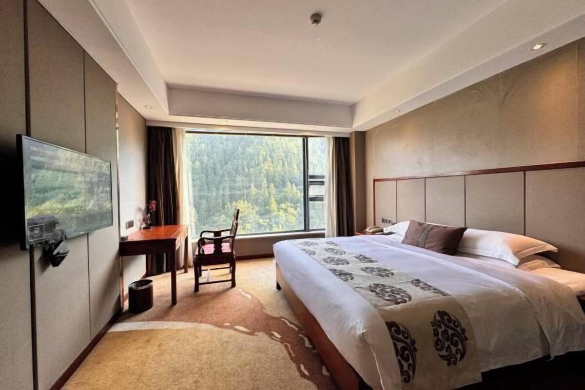 A hotel room with a large bed, a desk, a chair, a flat-screen TV, and a window with a forest view.