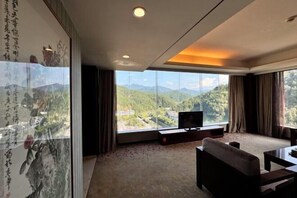A modern hotel room with a large window offering a mountain view, a flat-screen TV on a stand, and traditional artwork on the wall. - Ling Long Wang International Hotel (Guidong)