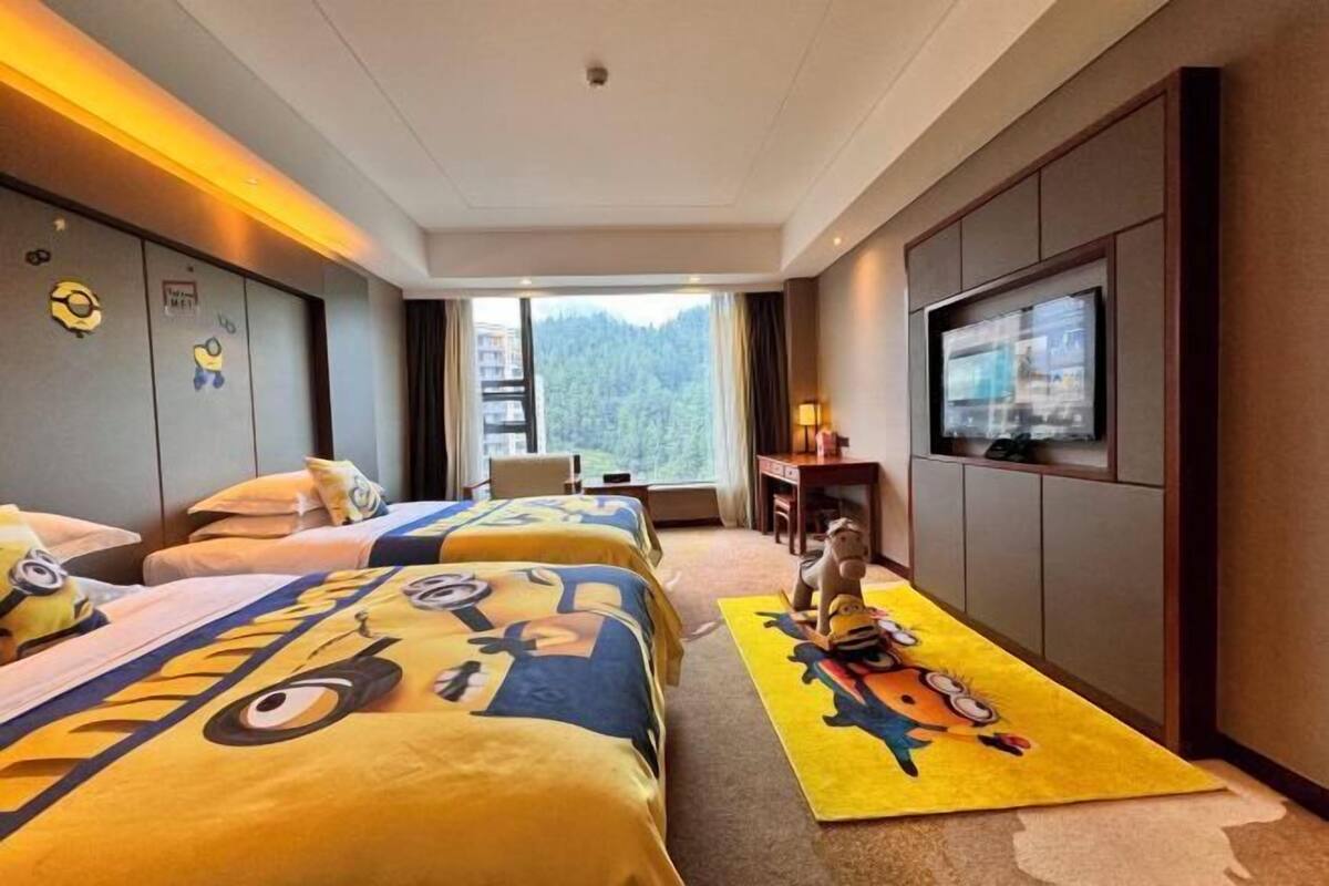 A hotel room with three beds, a TV, a desk, and a view of a forest.