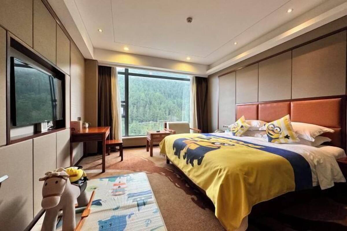 A hotel room with a large bed, a desk, a chair, a television, and a window with a forest view.
