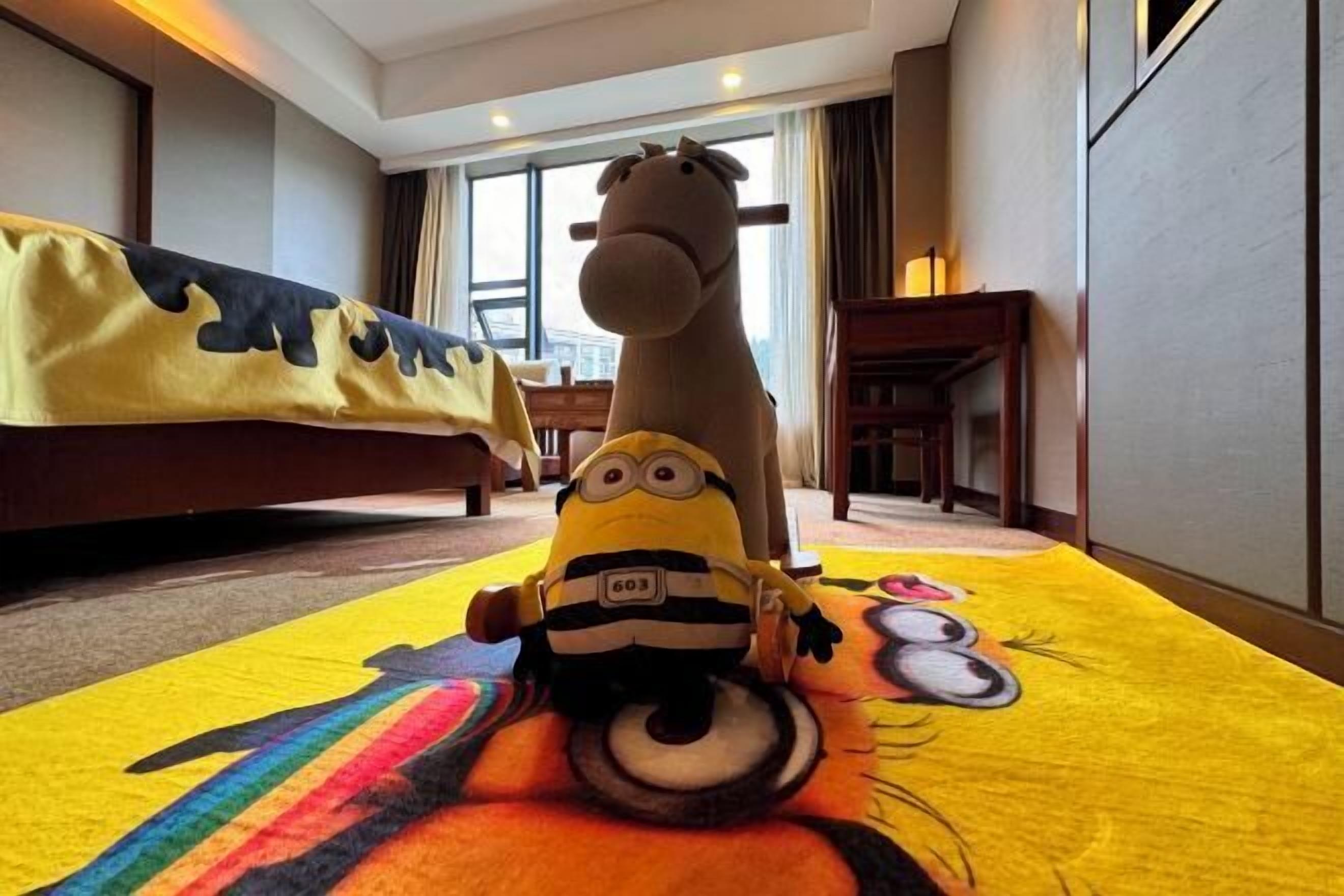 A hotel room with a large bed, a Minion plush toy on a colorful rug, and a giraffe statue.