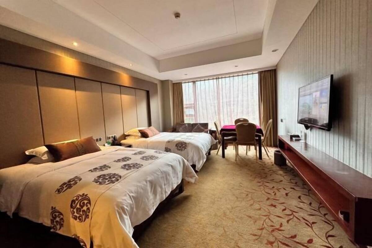 A hotel room with two beds, a desk, a chair, a TV, and a large window with curtains.
