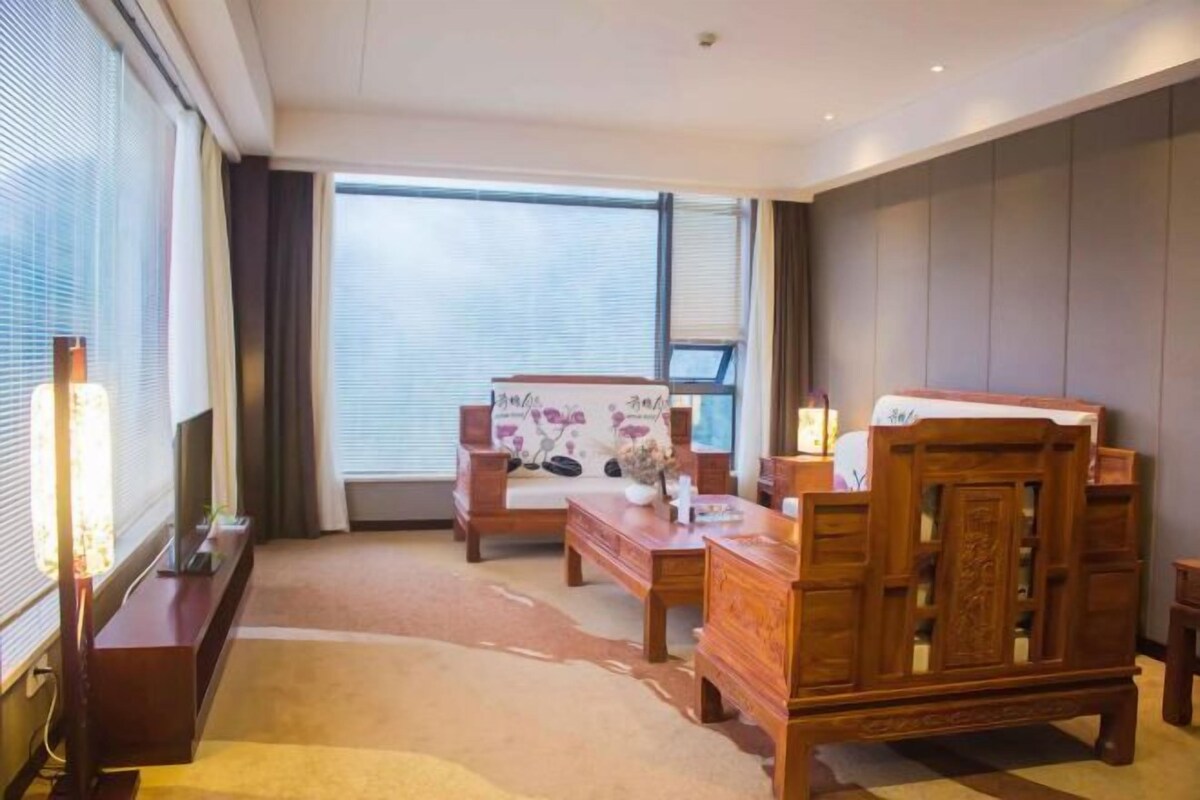 A hotel room with a large window, a wooden bed, a wooden dresser, a small table, and a TV on a stand.