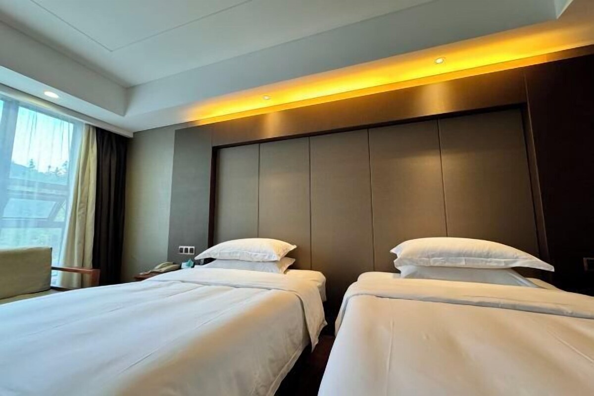 A hotel room with two beds, a large window with curtains, a bedside table with a lamp, and a headboard with built-in lighting.