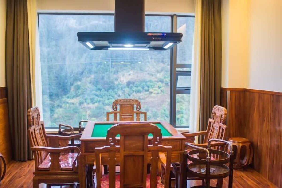 A billiards table with four chairs around it, a large window with a view of greenery, and a modern ceiling light fixture.