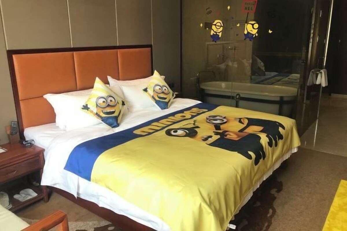 A hotel room with a bed featuring a Minions-themed bedspread, a wooden headboard, a bedside table with a lamp, and a bathroom visible through a glass door.