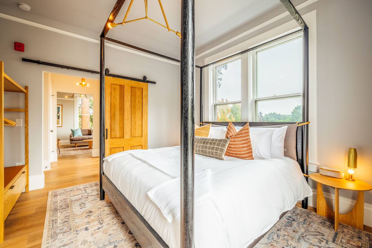 A bedroom with a four-poster bed, a wooden wardrobe, a bedside table with a lamp, and a large window.