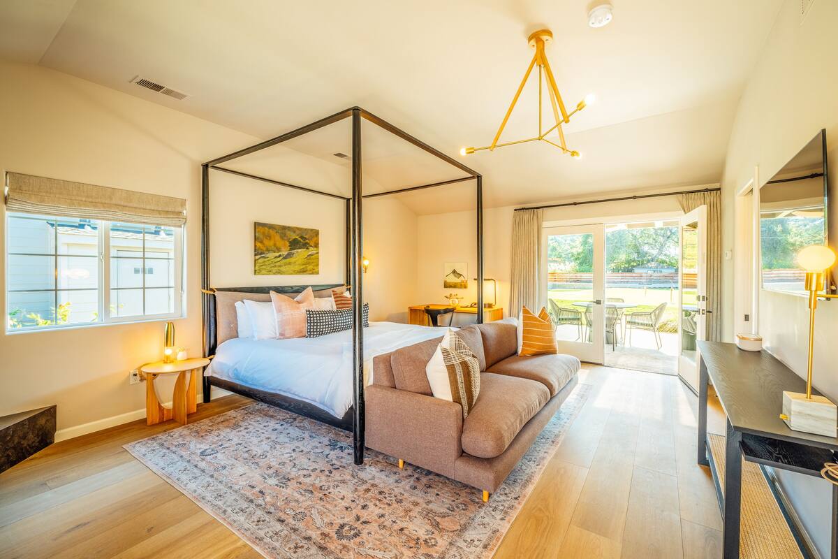 A bedroom with a four-poster bed, a sofa, a desk, and a large window with a view of the outdoors.