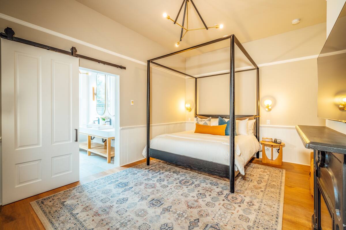 A bedroom with a four-poster bed, a wooden floor, a patterned rug, a bedside table with a lamp, and a sliding door leading to another room.
