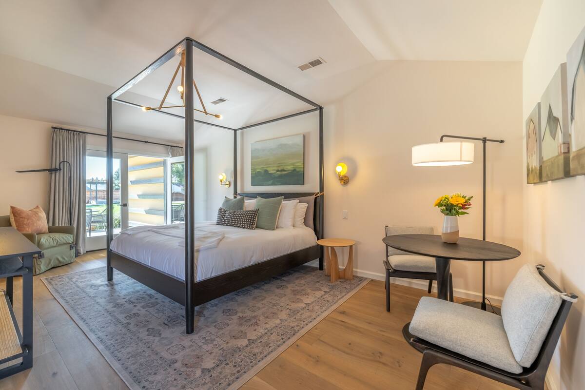 A modern bedroom with a four-poster bed, a round table, a chair, and a view of the outdoors.