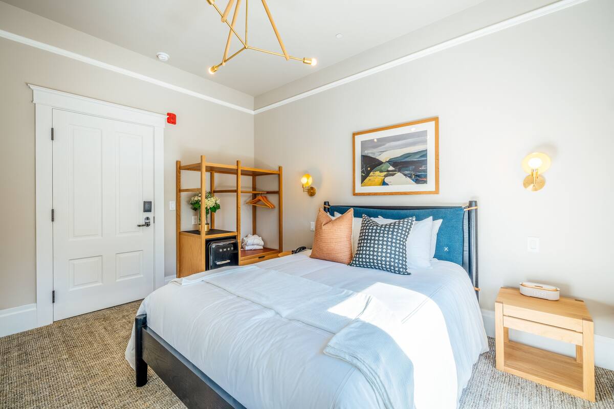 A neatly arranged bedroom with a bed, bedside table, and a wall-mounted shelf with towels and a plant.