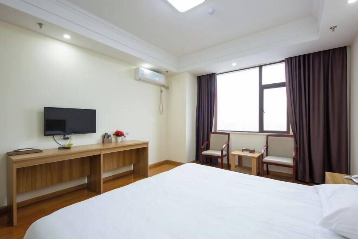 Ein Hotelzimmer mit einem großen Bett, einem Schreibtisch mit Fernseher, einem kleinen Tisch, zwei Stühlen und einem fenster mit Vorhängen.