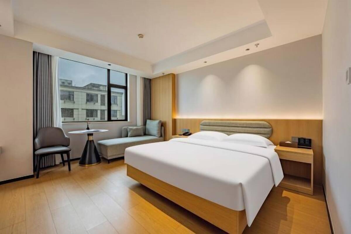 Ein modernes Hotelzimmer mit einem großen Bett, einem kleinen Tisch, einem Stuhl und einem Fenster mit Blick auf Gebäude.