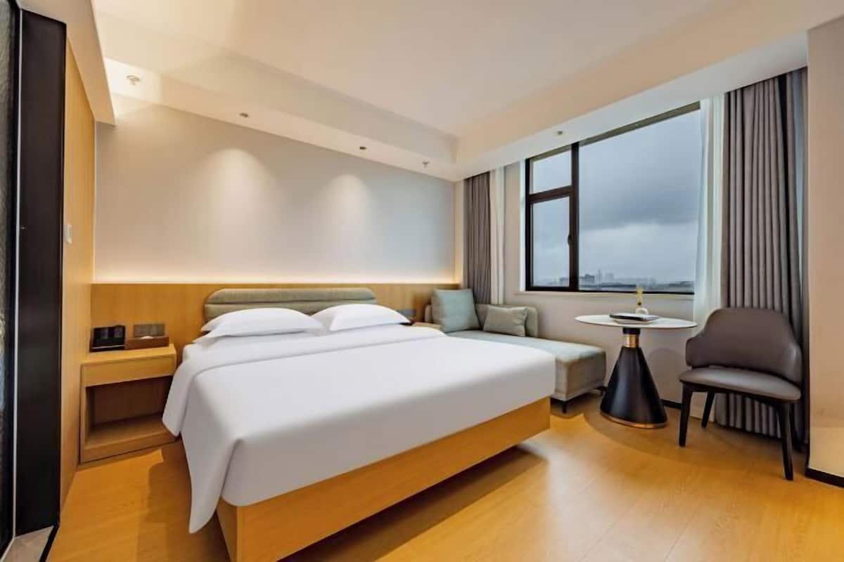 Ein modernes Hotelzimmer mit einem großen Bett, einer kleinen Sitzecke, einem runden Tisch und Blick auf die Stadt durch große Fenster.