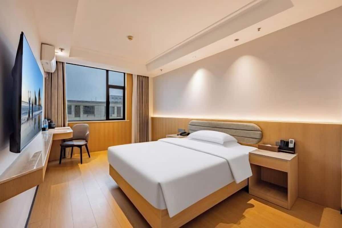 Ein Hotelzimmer mit einem großen Bett, einem Schreibtisch, einem Sessel, einem Fernseher und einem Fenster mit Blick auf die Stadt.
