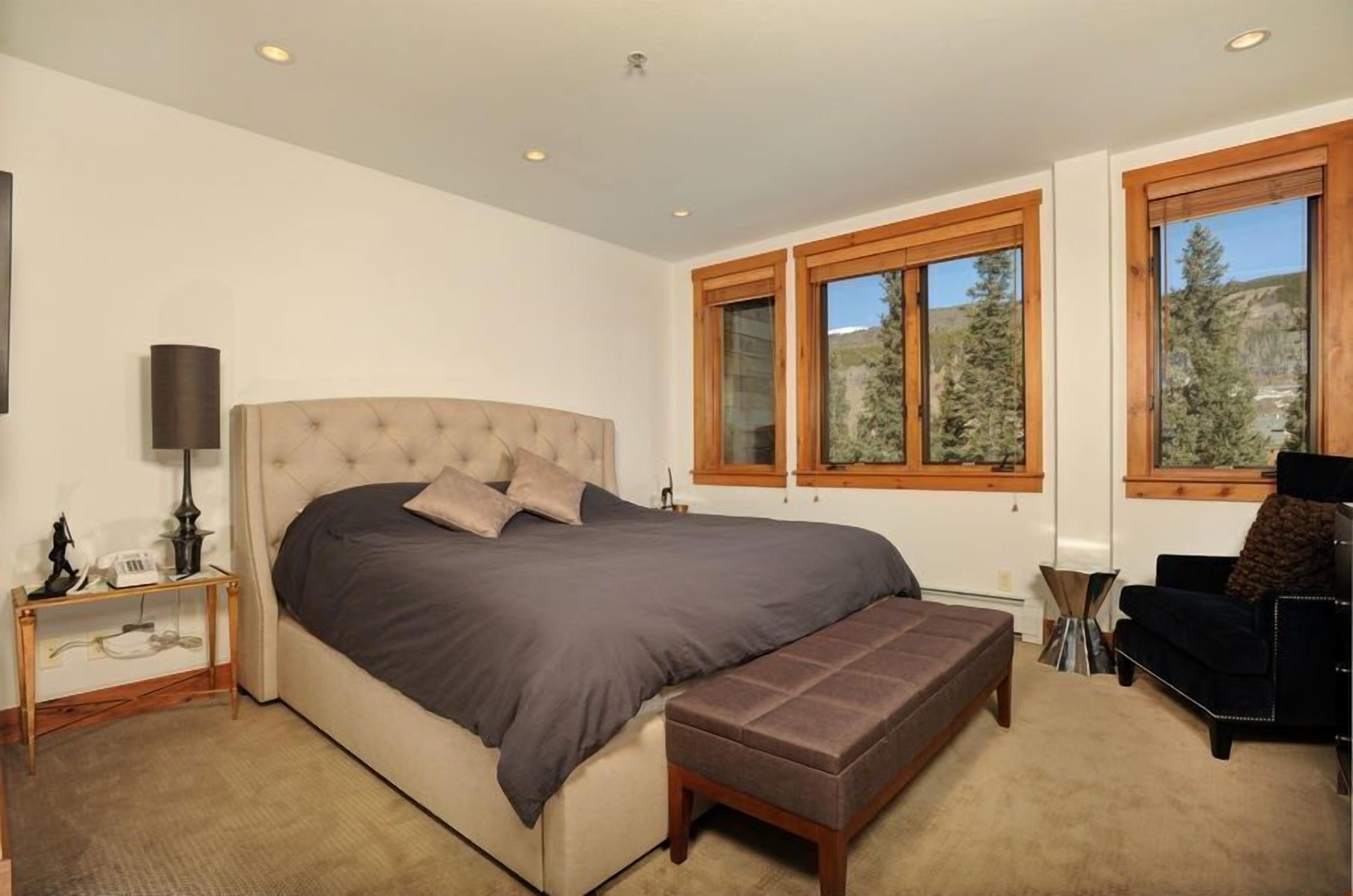 Un dormitorio con una cama grande, un banco, una silla y dos ventanas con vistas a árboles.