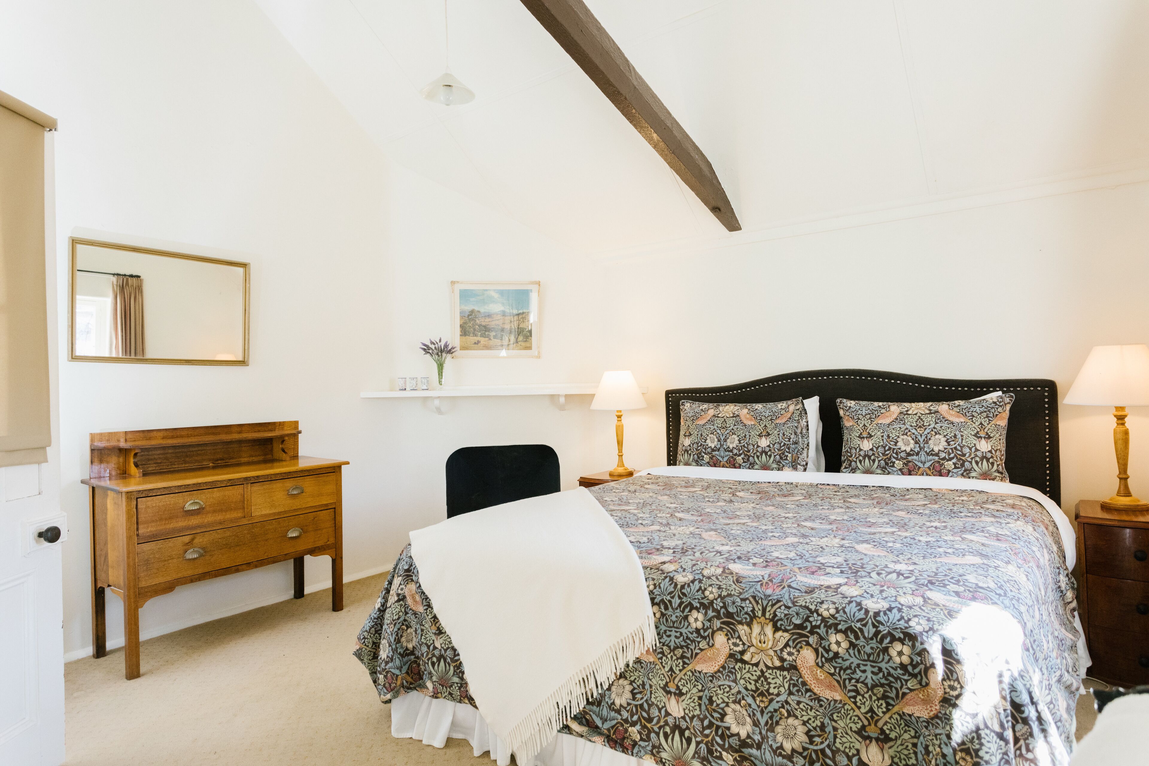 A bedroom with a bed, a wooden dresser, a chair, a nightstand with a lamp, a mirror, and a painting on the wall.