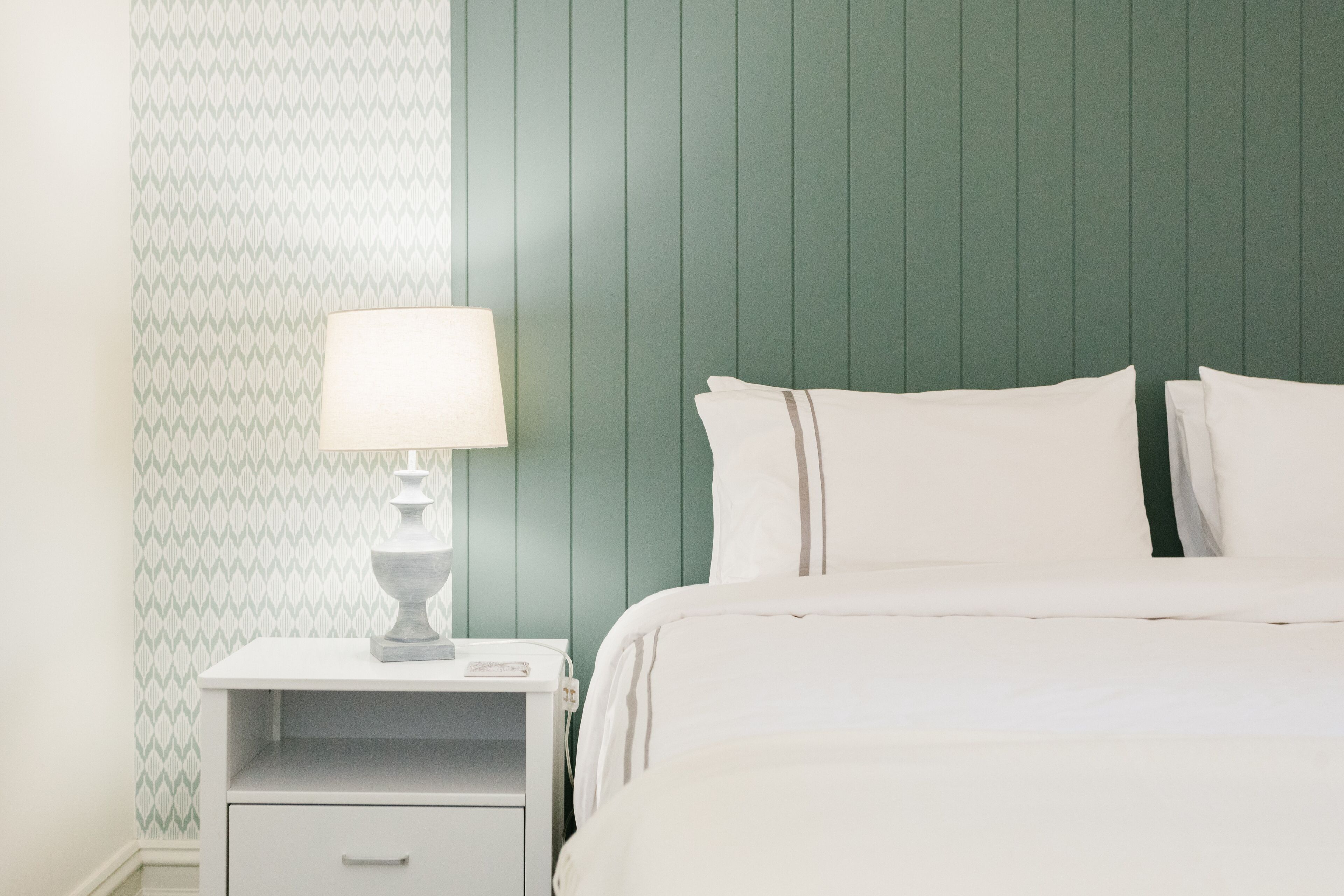 A neatly made bed with white linens and pillows, a bedside table with a lamp, and a patterned wallpaper on the left wall.