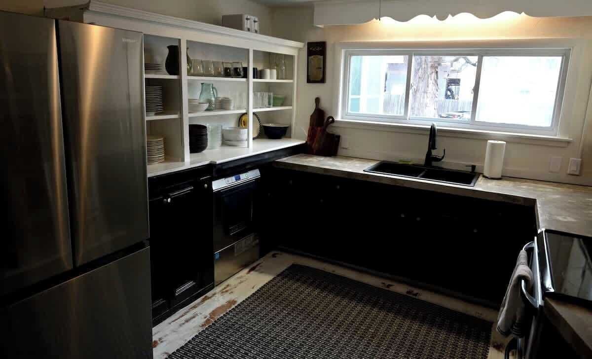 A kitchen with a stainless steel refrigerator, black cabinets, a window with a view, and a built-in shelf with dishes.