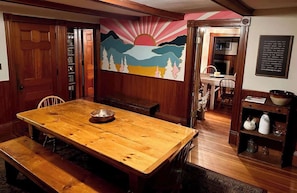 Una mesa de madera para comer con bancos, un mural de una puesta de sol y una estantería con objetos decorativos.