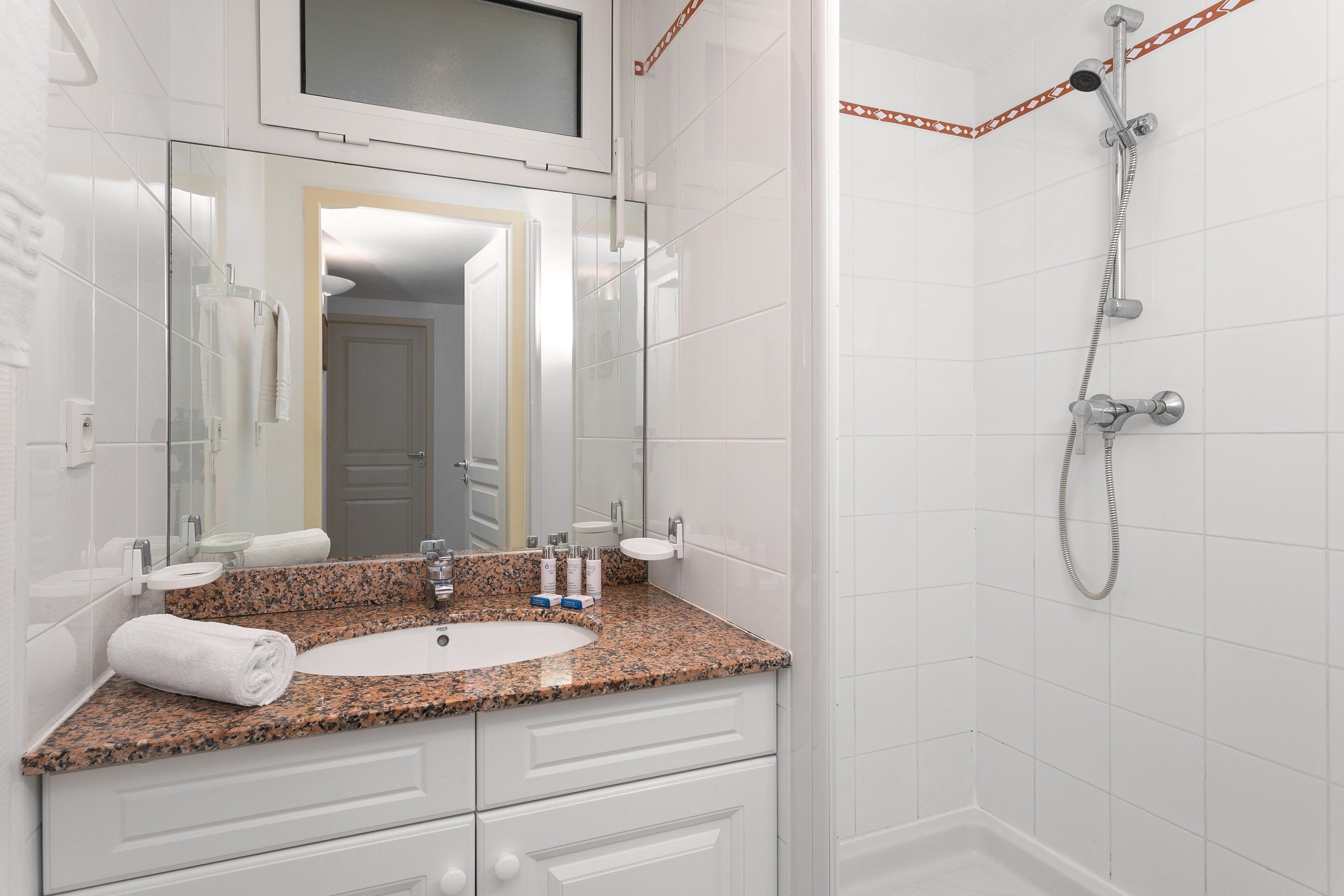 A bathroom with a granite countertop sink, a mirror, a shower with a handheld showerhead, and white tiled walls.