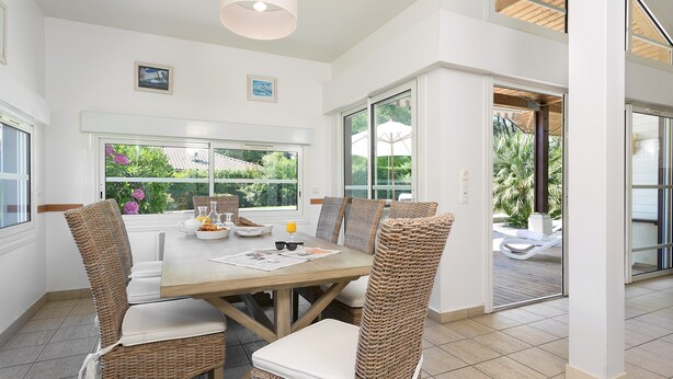 A dining area with a wooden table, wicker chairs, and a view of the garden through large windows. - Belle villa avec piscine privée | Près de la plage (Moliets)