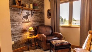 Una habitación acogedora con un sillón de cuero, una mesita pequeña, una estantería de madera con adornos y un tablero de ajedrez en la pared, además de una ventana con cortinas.