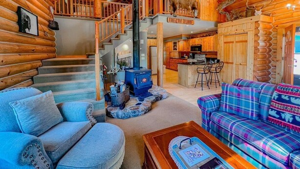 A log cabin interior with a staircase, a fireplace, a kitchen, and a living area with plaid furniture. - Bison Turf- Hot Tub-Views - Close to Downtown (Red Lodge)