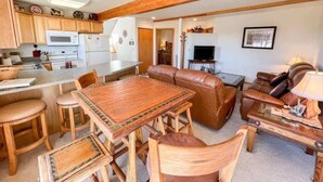 A cozy living area with a wooden dining table, leather sofa, and a kitchen with white appliances. - Black Bear Den- Mountain Views - Close to Downtown (Red Lodge)