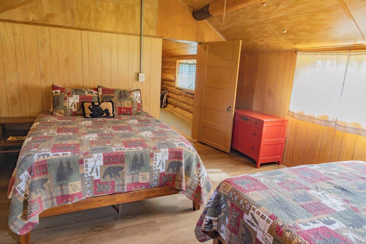 Um quarto de cabana aconchegante, com decoração em madeira, uma cama com colcha, uma cômoda vermelha e uma janela com cortinas.