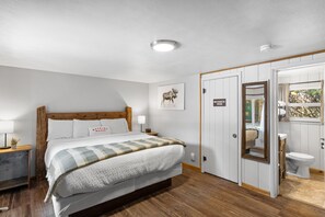 A bedroom with a wooden bed, a bedside table, a nightstand, a wall-mounted lamp, a framed picture, and a door leading to a bathroom. - The Bucking Moose (#08) Rabbit Room King Suite in West Yellowstone, 1 min to Yellowstone! (West Yellowstone)