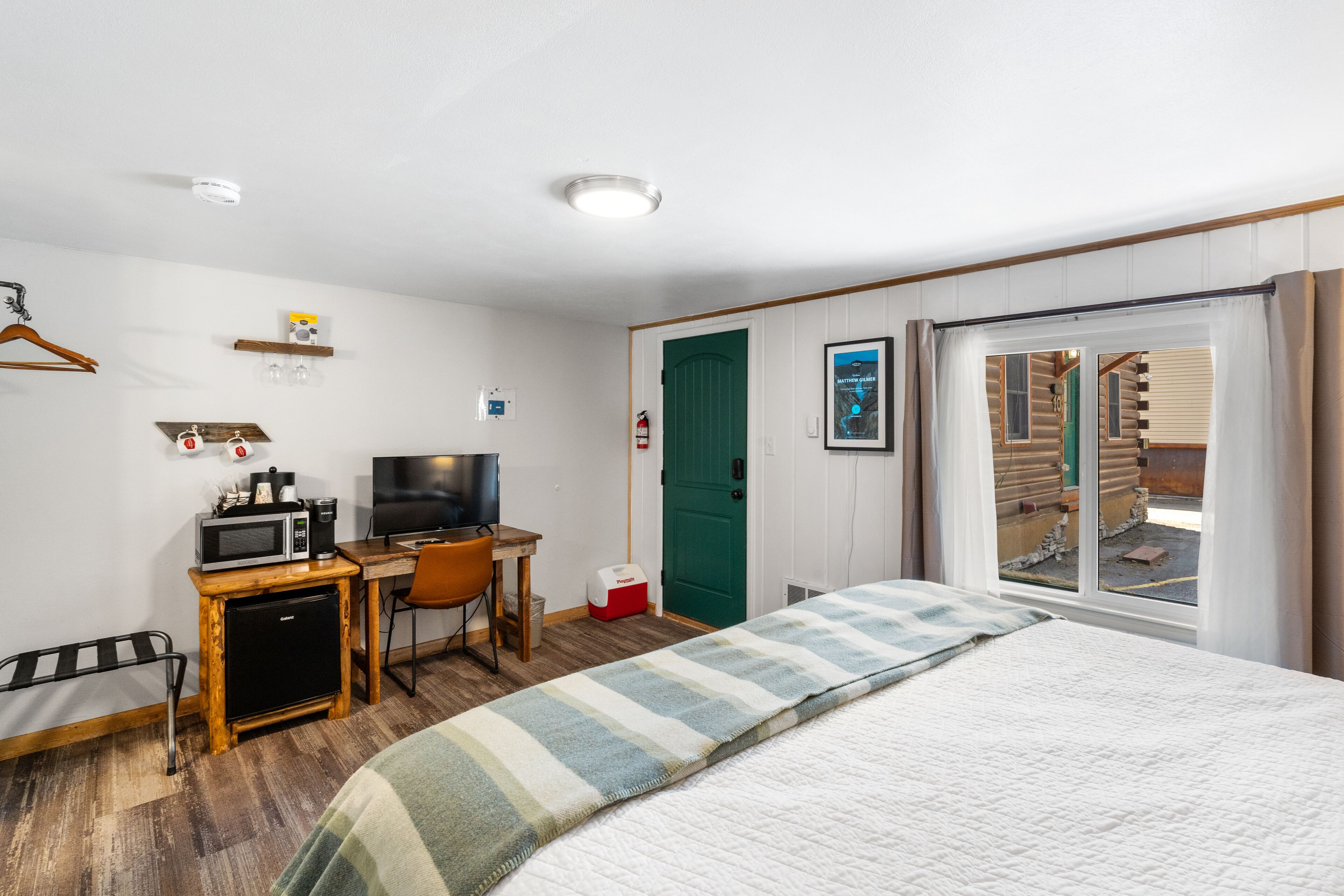 Habitación de hotel con cama, escritorio con televisor, microondas, refrigerador pequeño, silla, perchero y ventana con cortinas.