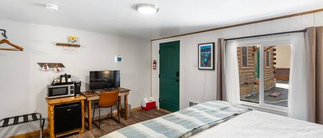 Habitación de hotel con cama, escritorio con televisor, microondas, refrigerador pequeño, silla, perchero y ventana con cortinas.