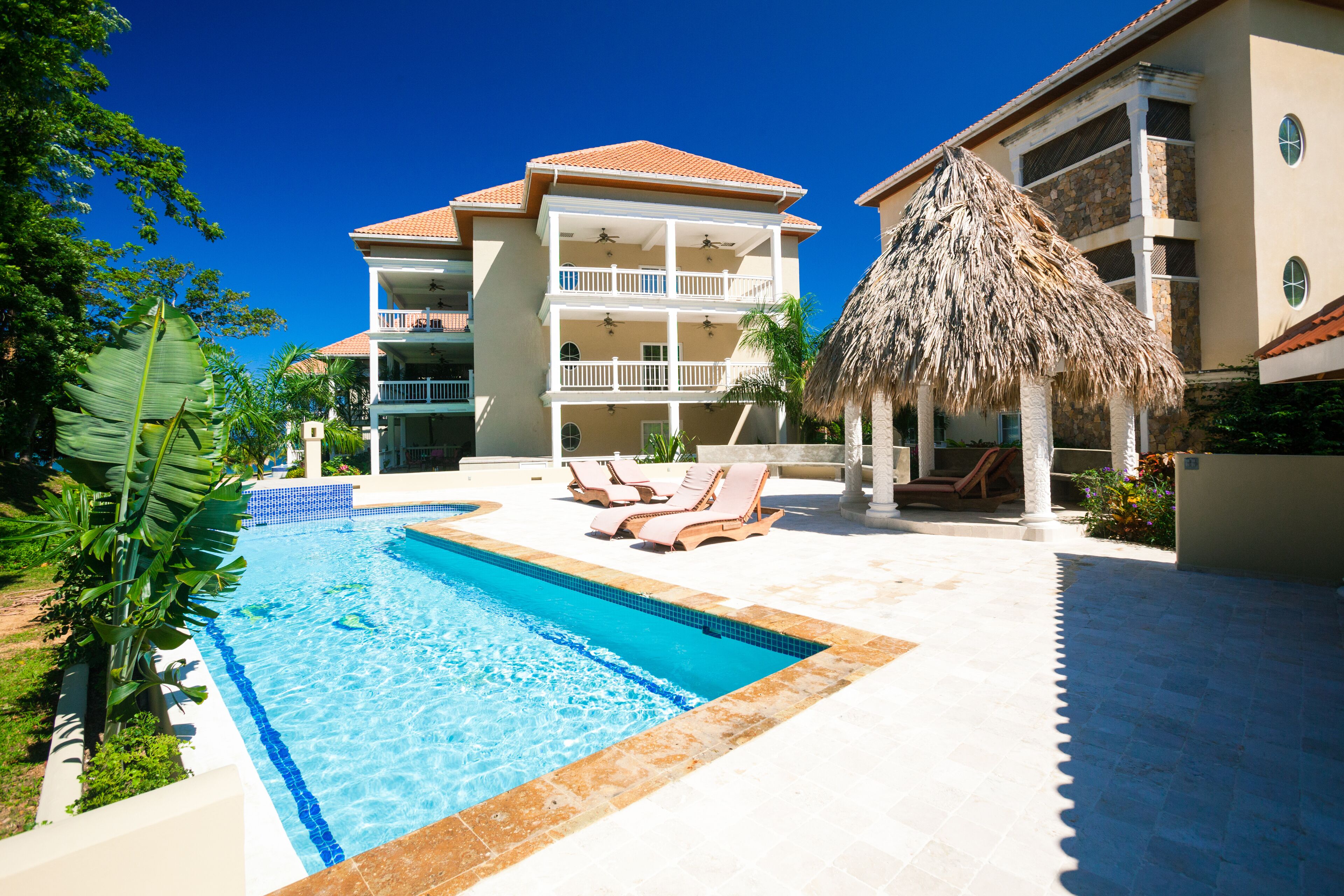 A swimming pool with lounge chairs, a thatched-roof structure, and a multi-story building with balconies.