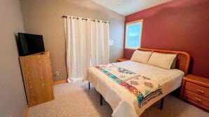 Une chambre à coucher comprenant un lit, une table de chevet, une armoire en bois, une fenêtre avec des rideaux et un luminaire au plafond.
