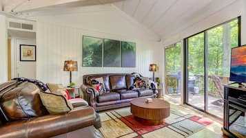 A spacious living room with a leather sofa, a coffee table, and a television. There are large windows offering a view of the outdoors.