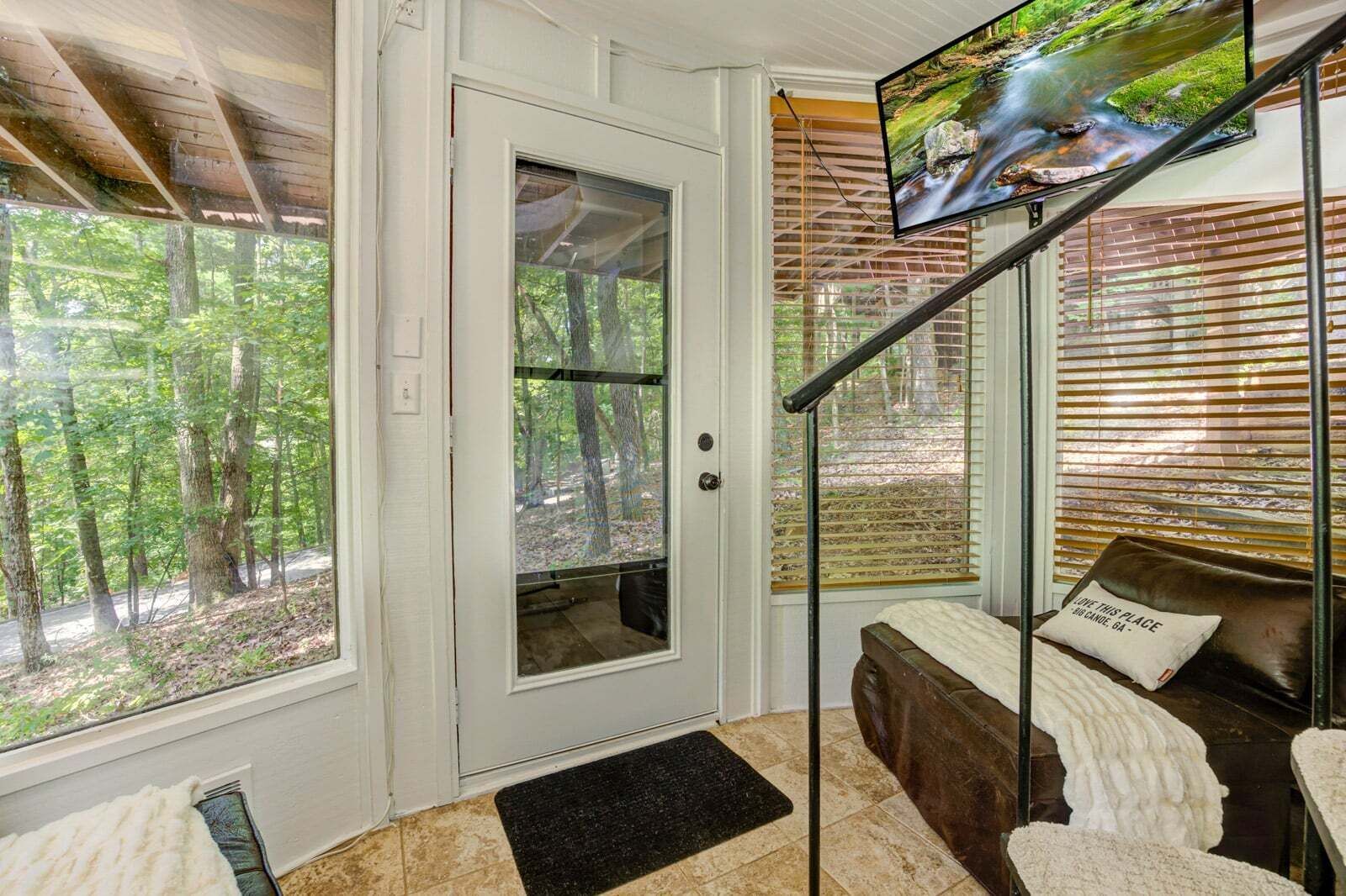 A room with a door leading to a forested area, a bed with a pillow, a TV on the wall, and a window with blinds.