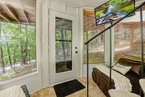 A room with a door leading to a forested area, a bed with a pillow, a TV on the wall, and a window with blinds.