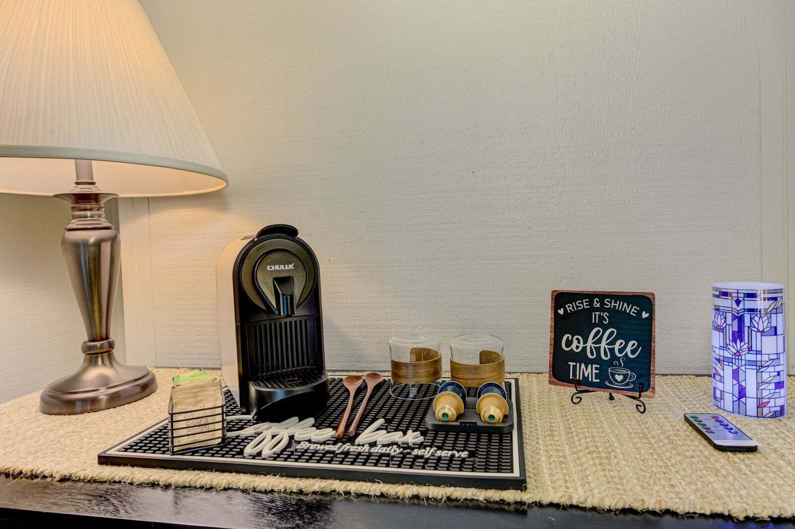 A coffee station with a coffee maker, cups, a sign, and a remote control on a textured mat.