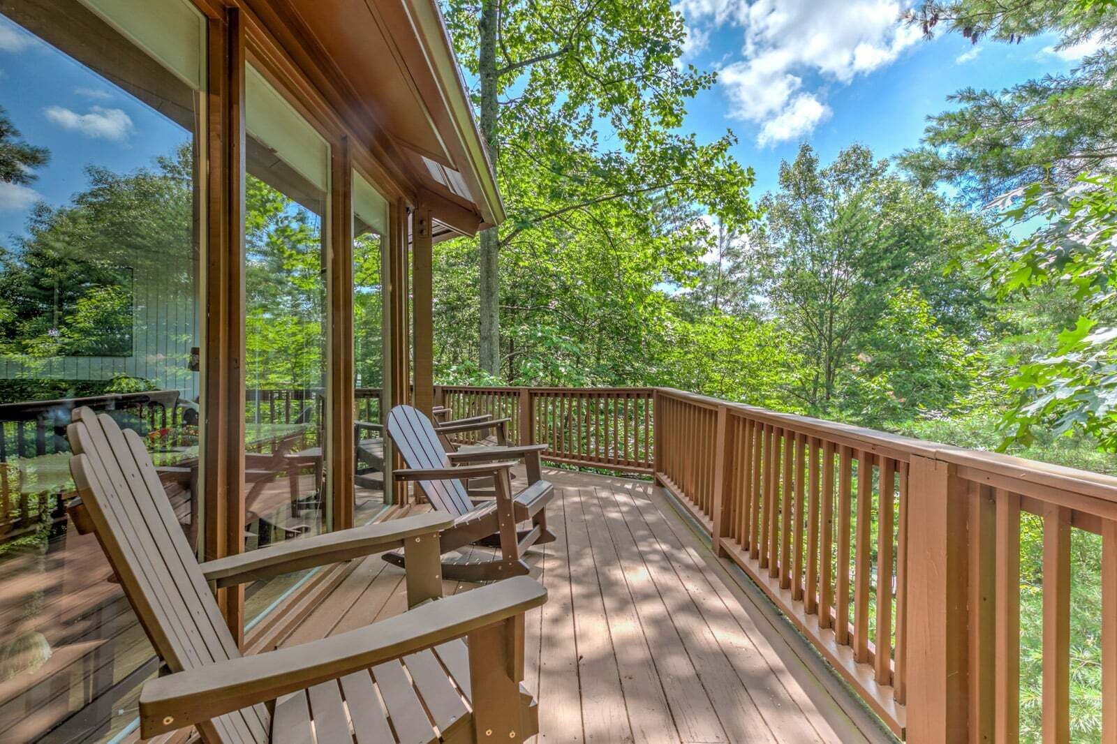 A wooden deck with Adirondack chairs, a glass enclosure, and a view of a forested area.