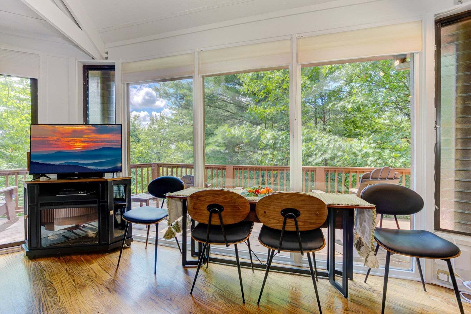 A sunroom with a TV, wooden chairs, a table, and a view of trees.