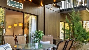 Un espace extérieur aménagé avec une table et des chaises en verre, une pancarte indiquant « Restez un peu », et une vue sur les arbres à travers les fenêtres.