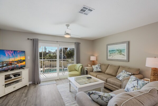 A living room with a TV, sofa, armchair, and a glass coffee table. There is a balcony with a pool view.