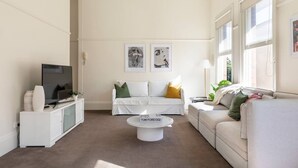 A modern living room with a white sofa, a coffee table, and a TV on a white cabinet. There are two framed pictures on the wall and a window with a view of greenery. - Oversized Artdeco Apartment w 4m high ceilings (Glebe)